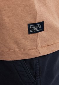 Krátký béžový bavlněný rukávový košile s námořnickou etiketou s nápisem „Petrol Ind“ a „Denim Driven Since ’93 Florida U.S.A.“, spárovaná s tmavými kalhotami.