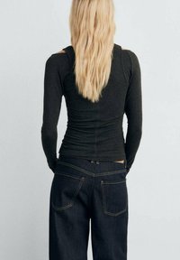 Haut noir à manches longues avec une silhouette ajustée, détails froncés à l'arrière, associé à un jean en denim bleu foncé avec des coutures et des poches visibles.