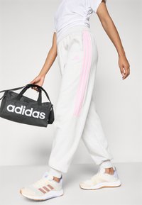 Person som bærer hvite Adidas joggebukser med rosa striper og beige Adidas sneakers, mens de holder en svart Adidas duffel bag på vei.