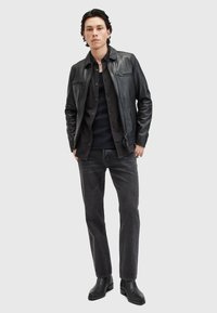 Zwarte leren jas over een zwart shirt, gecombineerd met donkergrijze jeans en zwarte boots. De jas heeft een ritszak en een kraag.