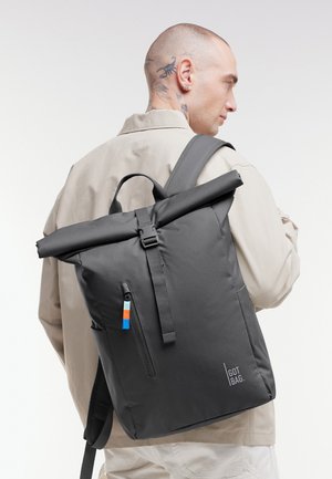 Hombre con la cabeza rapada y tatuajes en el cuello, vistiendo una chaqueta beige y llevando una mochila negra con tapa enrollable, con un bolsillo frontal con cremallera y tirador de cremallera colorido.