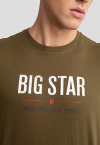 Kaki spalvos medvilninis marškinėliai su apvaliu kaklu, turintys tekstūruotą baltą "BIG STAR" grafiką ir atspausdintą tekstą raudona ir mėlyna spalvomis apačioje.