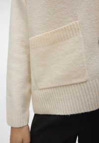 Cremefarvet sweater med ribbet kant og manchetter, der har en frontlomme. Teksturen ser blød og strikket ud.
