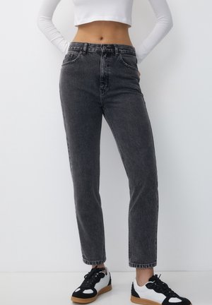 Frau trägt schwarze Jeans mit hoher Taille und Waschung, weißes, kurz geschnittenes Langarmshirt und schwarz-weiße Sneakers, steht vor einer schlichten weißen Wand.