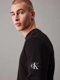 Crna teksturirana košulja dugih rukava s pletenim uzorkom, koja na lijevom rukavu ima pravokutnu etiketu s Calvin Klein Jeans logotipom.
