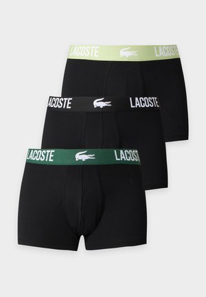 Мъж без риза, стоящ бос, носещ черни боксерки Lacoste с зелен колан и лого Lacoste на бял фон.