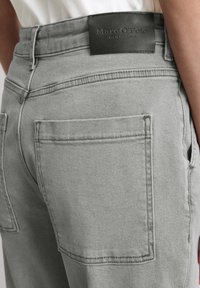 Pantalons en denim gris avec une coupe structurée, deux poches arrière et une étiquette en cuir à la taille. Texture lisse.