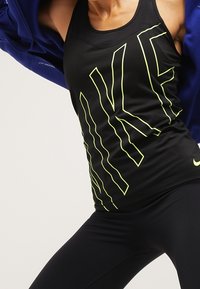 Černé atletické tílko s velkým geometrickým logem "NIKE" v zářivě žluté barvě. Design zahrnuje styl s racerbackem a těsný střih.