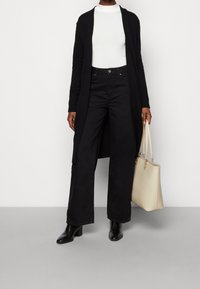 Lång svart cardigan över en vit polo, tillsammans med svarta vida byxor och svarta klackstövlar. Håller en beige handväska.