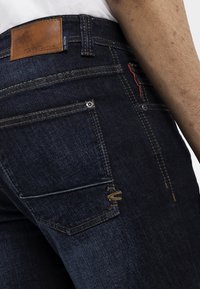 Zoom in de unos vaqueros de mezclilla azul oscuro con una etiqueta de cuero marrón de la marca y costuras visibles en el bolsillo trasero y la cinturilla.