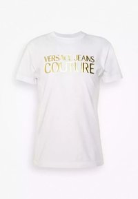 T-shirt bianca a maniche corte in cotone con collo rotondo. Presenta la scritta "VERSACE JEANS COUTURE" in lettere dorate lucenti sulla parte anteriore.