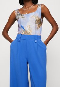 Topp: ärmlös, lätt tyg med blå himmel och cherubmönster. Botten: vida byxor i livfull blå med knapplösningar vid midjan.