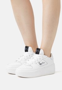 Valged sneakers, millel on tekstuuriga nahkpealts, nööridega disain, pehme krae ja must brändi silt. Omab paksu kummist välistalla.