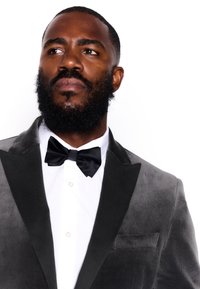 Veste de smoking en velours gris avec revers en satin noir, associée à une chemise blanche et un nœud papillon noir. Boutons visibles et détails de poche.