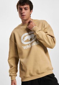 Sudadera beige con escote redondo y corte holgado. Presenta un gráfico de logo blanco de un rinoceronte y texto en el frontal. Puños y dobladillo acanalados.