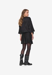 Top in satin nero con maniche ampie, abbinato a una gonna nera a campana. La modella indossa stivali neri al ginocchio con dettagli in fibbia.