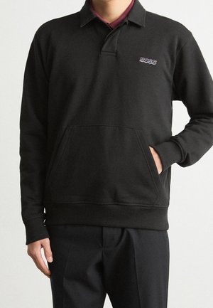 Mann trägt einen schwarzen Langarmpullover mit Kragen, Fronttasche und "BOSS"-Logo auf der Brust, kombiniert mit schwarzen Hosen.