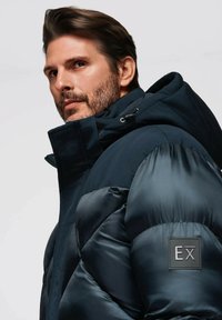 Mörkblå puffjacka med hög krage, med en glänsande yta, dragkedjestängning och en logotypapplikation på vänster ärm.