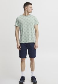 Grønn t-skjorte med korte ermer og sirkulære mønstre, kombinert med marineblå shorts. Modellen har på seg hvite ankelstrømper og mørke joggesko.