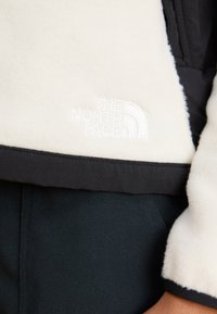 Bunda z fleece v krémovej a čiernej farbe s vyšitým logom; má jemnú textúru a kontrastný tmavý lem. Detailný pohľad, minimalistický dizajn.