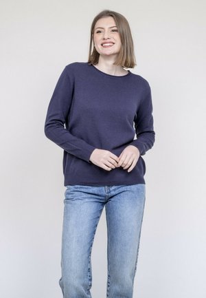 Jeune femme souriante portant un pull bleu marine et un jean bleu clair, debout devant un fond clair uni.