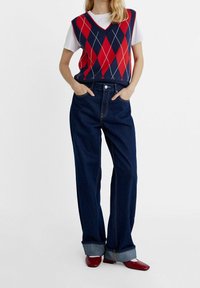 Persoon in een mouwloze rood en marineblauwe ruitjes sweater vest over een witte t-shirt, donkerblauwe omgeslagen wijde jeans, en rode vierkante schoenen.