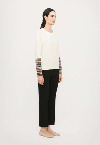Pullover crema con maniche a righe in vari colori, abbinato a pantaloni neri e ballerine beige. Tessuto morbido, vestibilità rilassata e design semplice.