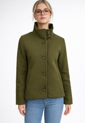 Vrouw draagt een olijfgroene jas met knopen en hoge kraag, en lichtblauwe spijkerbroek, staand tegen een witte achtergrond.