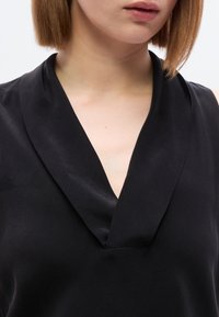 Μαύρο αμάνικο τοπ με βαθύ V-λαιμόκοψη και απαλή, μεταξωτή υφή. Το ύφασμα έχει διακριτική λάμψη και είναι ραμμένο για εφαρμογή που αγκαλιάζει το σώμα.