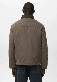 Giacca marrone robusta con colletto strutturato, cuciture visibili e silhouette squadrata, con maniche lunghe e orlo leggermente curvo.