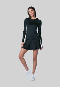 Zwart sportshirt met lange mouwen, ribbelstructuur en witte accenten aan de zijkanten, gecombineerd met een zwarte geplooide rok. Model draagt witte sneakers.