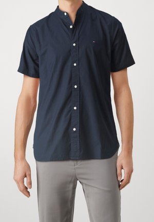 Homme portant une chemise à manches courtes bleu marine à boutons avec un col mao et un pantalon gris, sur un fond uni.