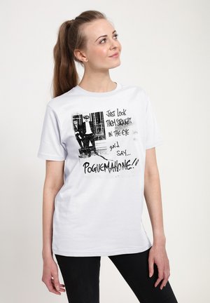 Femme aux longs cheveux en queue de cheval porte un t-shirt blanc avec un graphisme noir représentant deux hommes et un texte disant "Just look them straight in the eye and say... Poguemaione."