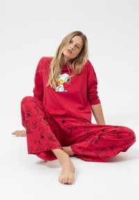 Vrouw met lang blond haar die blootsvoets op de vloer zit en een rode lange-sleeve top met Snoopy-thema en bijpassende broek met Peanuts-figuren draagt.