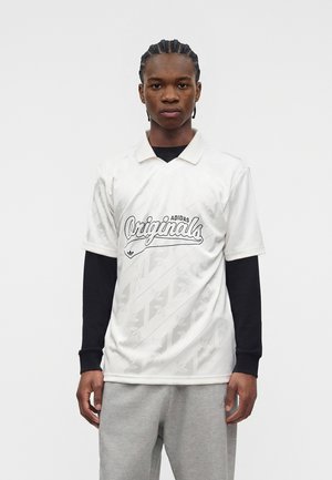 Mladý muž s pletenými vlasmi, ktorý má na sebe biele polo Adidas Originals cez čierne tričko s dlhým rukávom a sivé tepláky, pozerá dopredu.
