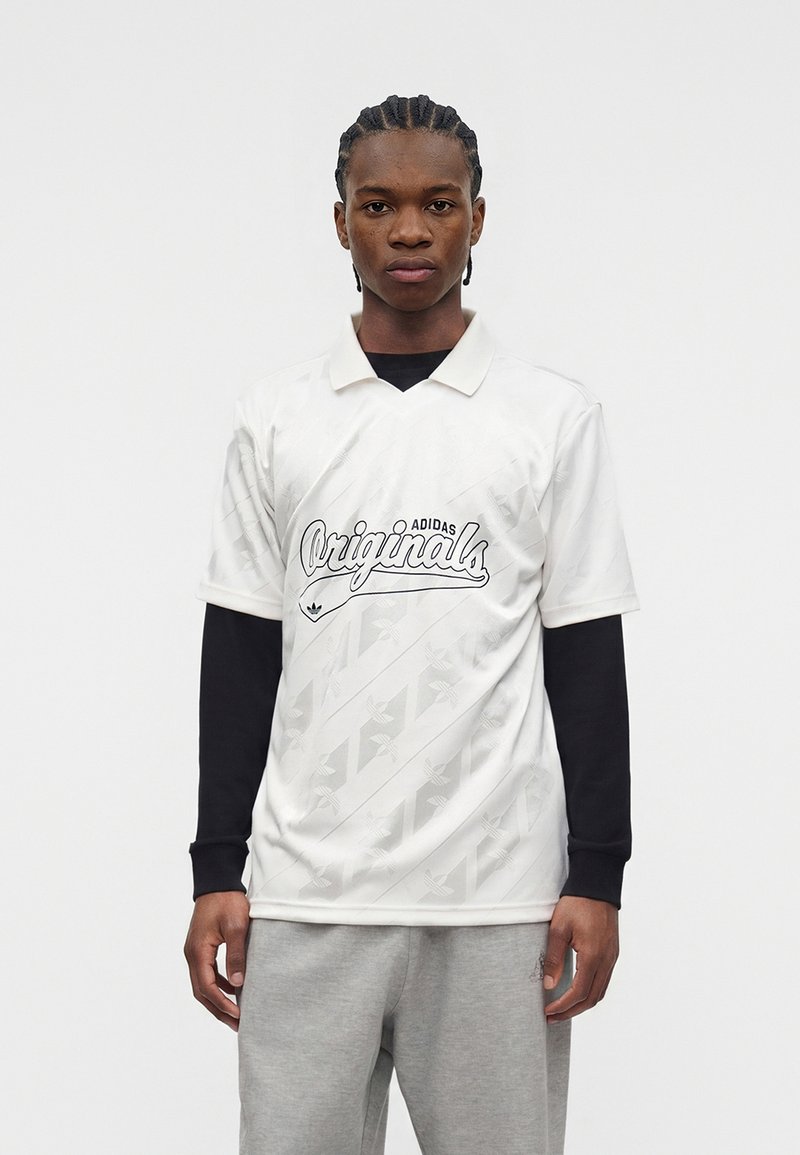 Νέος άνδρας με πλεξούδες, φοράει λευκό πόλο Adidas Originals πάνω από μαύρο μακρυμάνικο και γκρι φόρμα, κοιτάζοντας προς τα εμπρός.