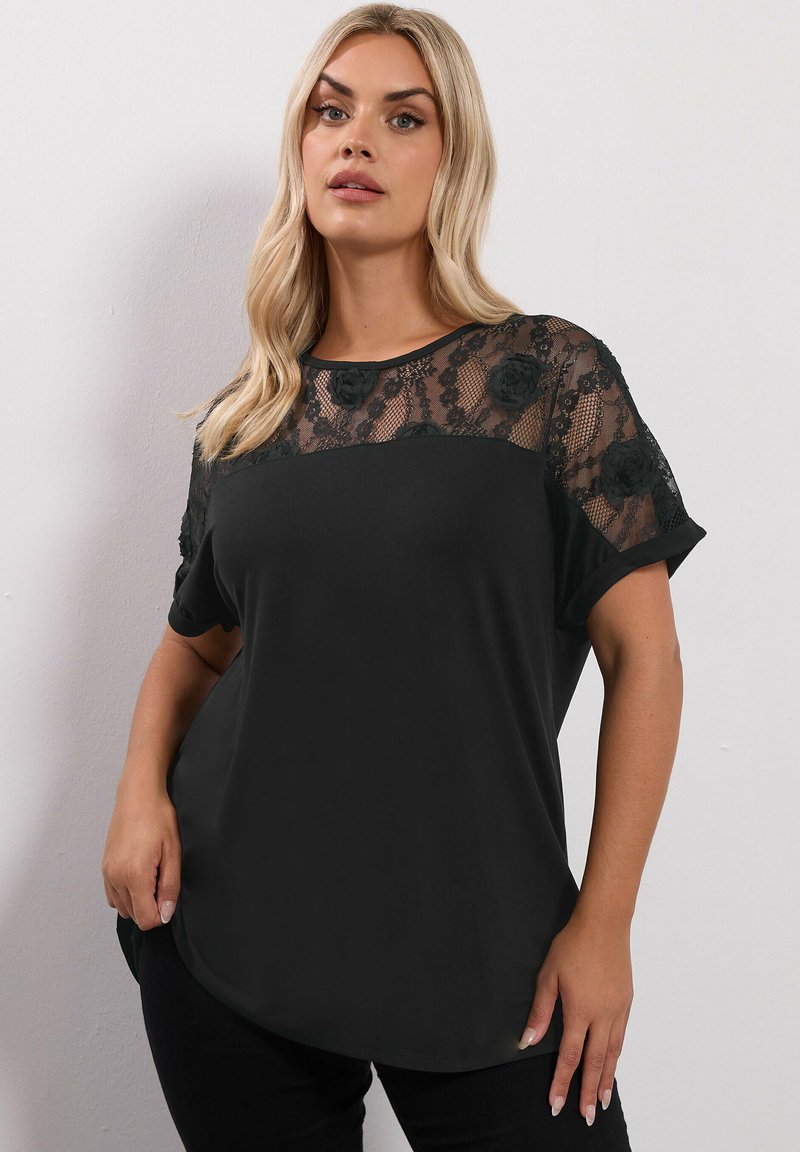 Top corto nero a maniche corte con yoke in pizzo e appliqué floreali. Corpo in tessuto liscio, orlo arrotondato e vestibilità comoda.