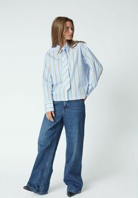 Blau-weiß gestreiftes, übergroßes Hemd mit Kragen, kombiniert mit weit geschnittenen blauen Jeans, akzentuiert durch spitze schwarze Schuhe.