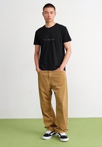 Svart T-skjorte med trykt logo, kombinert med lysebrune cord-bukser og svarte joggesko med hvite detaljer, mot en lys bakgrunn.
