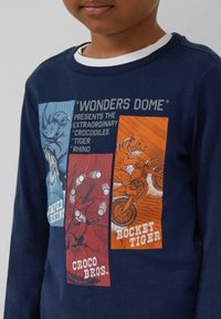 Kind draagt een marineblauwe sweatshirt met illustraties van een neushoorn, een juggelende krokodil en een tijger die op een motorfiets rijdt, met de teksten "Wonders Dome," "CROCO BROS," en "ROCKET TIGER."