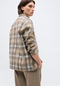 Άτομο με μεσαίου μήκους μαλλιά που φοράει πουκάμισο με μπεζ και λευκό καρό και καφέ παντελόνι, στέκεται με το ένα χέρι στην τσέπη, κοιτάζοντας πάνω από τον ώμο.