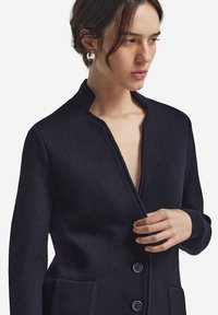 Granatowy sweter z fakturowanej tkaniny, z głębokim dekoltem w kształcie V, z zapięciem na guziki z przodu i dwoma przednimi kieszeniami. Posiada gładki splot i dopasowany krój.