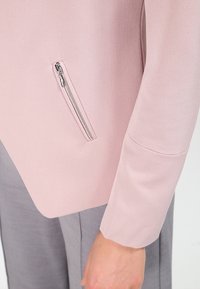 Veste rose clair à la texture lisse, avec une poche zippée argentée sur le côté gauche et une coupe ajustée. Complétée par un pantalon gris.
