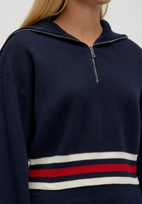 Marineblauer Reißverschluss-Hoodie aus strukturiertem Material. Verfügt über einen gestreiften Bund in Weiß, Marineblau und Rot. Enthält eine Kapuze und einen sichtbaren Reißverschluss.