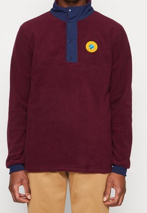 Mann trägt einen burgunderroten Fleecepullover mit marineblauem, geknöpftem Kragen und rundem gelbem Logo-Patch auf der Brust, kombiniert mit beigen Hosen.