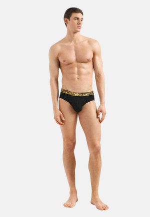 Giovane uomo in piedi a piedi nudi con slip neri con una fascia in vita dorata con testo ripetuto, su uno sfondo bianco semplice.