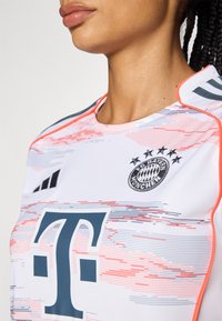 Maglia da calcio del Bayern Monaco con una base bianca decorata da motivi rosa e grigi, grande logo blu e badge del club ricamato sul petto.
