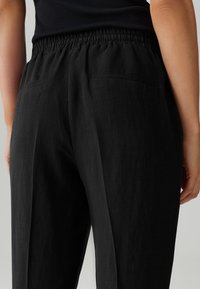 Pantalons en lin noir avec une taille élastique, dotés de deux poches arrière et d'une coupe décontractée, mettant en valeur un tissu texturé.