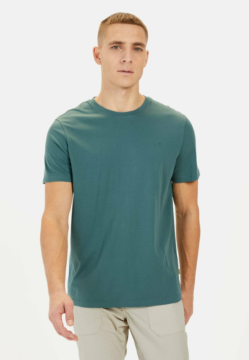 Teal crew-neck T-shirt lavet af blødt bomuld med korte ærmer og et lille logo på brystet. Modellen har lyse bukser på.