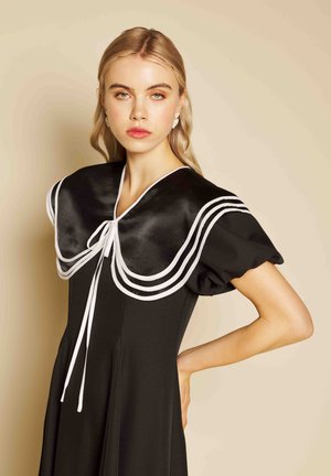 Jonge vrouw met blond haar draagt een zwarte jurk met grote matrozenkraag afgezet met wit, poseert met hand in de zij tegen een beige achtergrond.